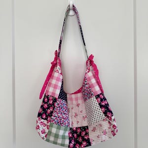May include: A patchwork shoulder bag with a variety of floral, polka dot, and gingham patterns in pink, navy, and green. The bag has a curved shape and a long strap with pink bows at the top.