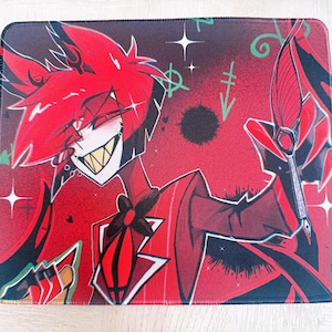 May include: Rectangular mouse mat featuring a cartoon character with red hair, a white face, and a wide grin. The character is set against a red background with black accents and green symbols. The mouse mat is designed for computer use.