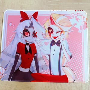 May include: A rectangular mouse mat with a vibrant anime-style illustration. Two characters are shown, one with long white hair and a red outfit, the other with blonde hair and a white suit. The background is pink with heart shapes.