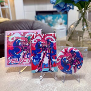 May include: Three items featuring a colourful anime-style character: a square print with the word "VELVET", a rectangular print, and a round button. The prints and button are in clear plastic sleeves and displayed on small clear stands. The character has pink and blue hair and clothing.