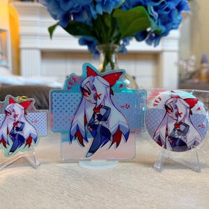 May include: Three acrylic character displays and a button. The character has white hair, a red bow, and a dark outfit. The displays are on clear stands. A bouquet of blue flowers is in the background.