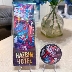 May include: A bookmark and a round pin featuring colorful artwork from "Hazbin Hotel." The bookmark is rectangular with the show's title at the bottom. The pin has a black border and a similar design. Both are displayed on clear stands.