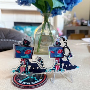 Może przedstawiać: Dwie akrylowe figurki przedstawiające postać z kreskówki z głową w kształcie ekranu telewizora, czerwonymi oczami i muszką. Jedna to brelok, druga stoi na podstawce z napisem "NO WIFI SIGNAL". Postać trzyma popcorn.