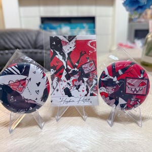 May include: A set of merchandise featuring characters from Hazbin Hotel. The set includes two round pinback buttons and a rectangular card. The buttons and card feature red, black, and white illustrations of characters.