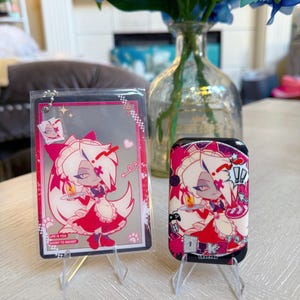 May include: Two items with anime-style artwork. One is a clear acrylic keychain with a pink and white character design and the text "LIFE IS TOO SHORT TO REGRET". The other is a black phone case with a similar design.