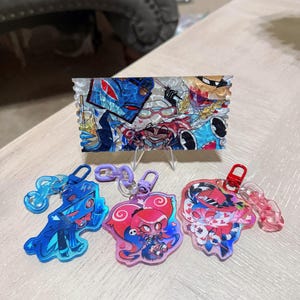 Może przedstawiać: Kolekcja kolorowych breloków akrylowych i panel dekoracyjny z postaciami z kreskówek. Breloki mają różne kształty i kolory, w tym niebieski, różowy i czerwony. Panel ma wielokolorowy wzór.