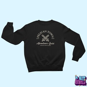 K&ouml;nnte beinhalten: Schwarzes Sweatshirt mit wei&szlig;em Aufdruck. Der Aufdruck zeigt den Text "Pelican Town" in einem kreisf&ouml;rmigen Design mit gekreuzten Schwertern und den Worten "Adventurer's Guild" und "Strive for Glory". Das Sweatshirt hat lange &Auml;rmel und einen Rundhalsausschnitt.