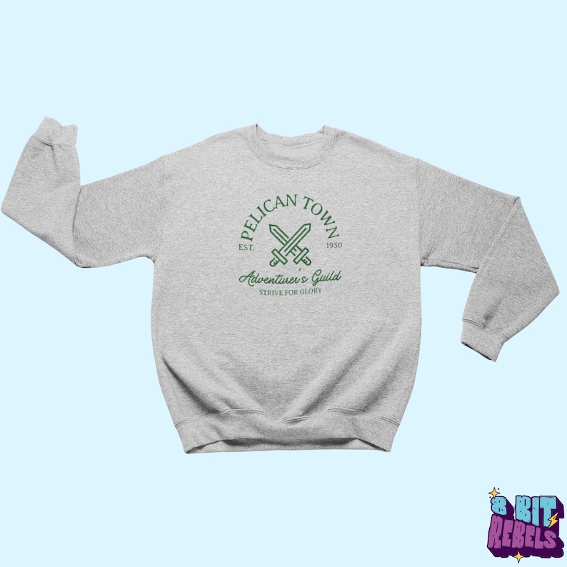 K&ouml;nnte beinhalten: Ein hellgraues Sweatshirt mit Rundhalsausschnitt und langen &Auml;rmeln. Auf der Vorderseite befindet sich eine gr&uuml;ne, kreisf&ouml;rmige Grafik mit dem Text "Pelican Town" und "Adventurer's Guild" sowie gekreuzten Schwertern. Darunter steht der Text "Strive for Glory".