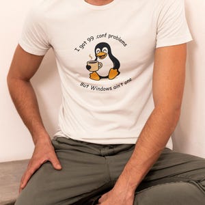 May include: White t-shirt featuring a cartoon penguin holding a coffee cup. The text on the shirt says "I got 99 .conf problems But Windows ain't one". The penguin is black and white with yellow accents. The person is wearing green trousers.