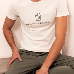May include: White crew neck t-shirt with a black graphic of a bin and the text "sudo rm -rf / bad vibes". The person is wearing olive green trousers and black trainers.