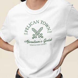 K&ouml;nnte beinhalten: Wei&szlig;es T-Shirt mit einem gr&uuml;nen Aufdruck mit dem Text "PELICAN TOWN" &uuml;ber gekreuzten Schwertern und "Adventurer's Guild" darunter. Das Shirt enth&auml;lt auch den Text "EST. 1950" und "STRIVE FOR GLORY".