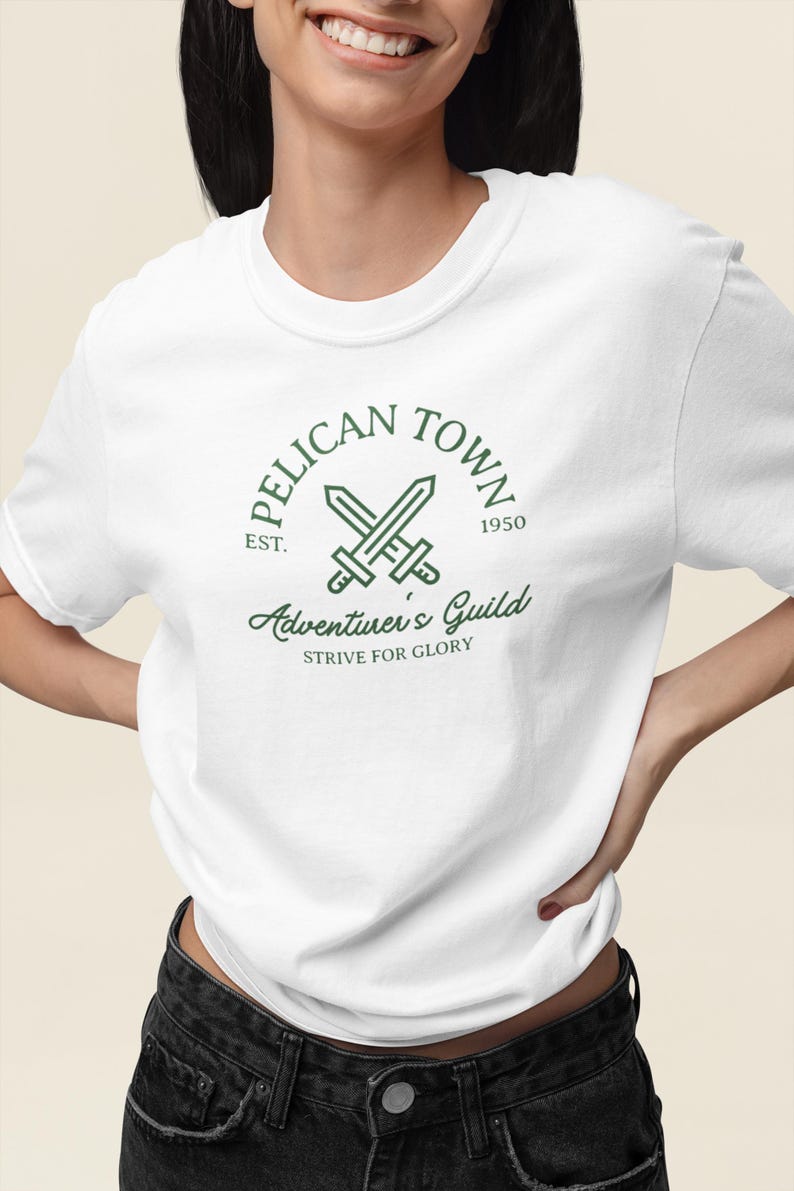 K&ouml;nnte beinhalten: Wei&szlig;es T-Shirt mit einem gr&uuml;nen Aufdruck mit dem Text "PELICAN TOWN" &uuml;ber gekreuzten Schwertern und "Adventurer's Guild" darunter. Das Shirt enth&auml;lt auch den Text "EST. 1950" und "STRIVE FOR GLORY".