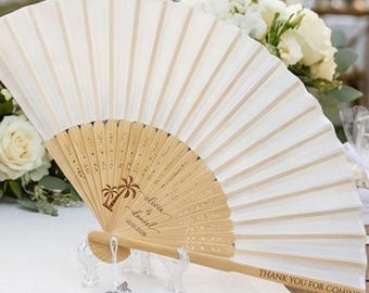 May include: Two off-white folding hand fans with light wood ribs, each featuring a white tassel at the base. The fans are printed with custom text including names, dates, and the phrase Thank You For Coming, displayed on a white table runner.