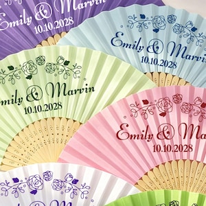 May include: A collection of six folding hand fans in purple, light blue, light green, pink, white, and bright green. Each fan features a wooden frame and is printed with the text Emily & Marvin, the date 10.10.2028, and a floral rose graphic.