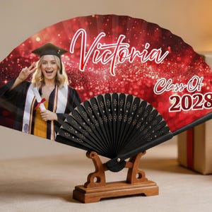 May include: A personalized graduation fan featuring a photo of a graduate in a cap and gown. The fan has the name "Victoria" and "Class of 2028" in red script. The fan is black and is displayed on a wooden stand.