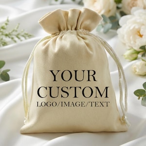 May include: A beige drawstring bag with the text "YOUR CUSTOM LOGO/IMAGE/TEXT" in black. The bag is made of a soft material and has a drawstring closure. The background features white fabric and flowers.
