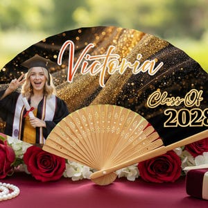 May include: A graduation fan with the name "Victoria" in red script and "Class of 2028" in gold. The fan features a photo of a graduate in a cap and gown. The fan is made of wood and has a decorative design.