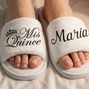 以下が含まれることがあります： 黒いスクリプトで「Mis Quince」と「Maria」の文字が入った白いプラッシュスリッパ。スリッパには「Mis Quince」という言葉の上に王冠のグラフィックがあります。
