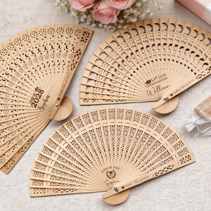 May include: Three intricately carved wooden fans with decorative cut-out patterns. Each fan features personalized text, including names and the year "2028", suggesting a graduation theme. The fans are light brown in color and have a natural wood finish.