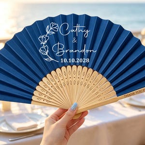 May include: A navy blue folding hand fan with light wood ribs. The fan features white printed text that reads Cathy & Brandon 10.10.2028 with a floral wreath graphic. The right wooden rib is engraved with the text THANK YOU FOR COMING.