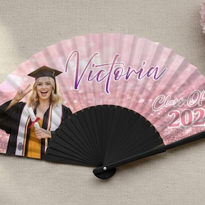 Puede incluir: Abanico de graduación personalizado, rosa, con foto de graduada con toga y birrete. El abanico tiene impreso el nombre "Victoria" y "Class of 2028". El abanico tiene un mango negro.