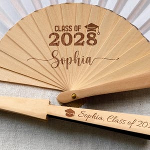Op de afbeelding: Een lichtbruine houten waaier met de tekst "CLASS OF 2028 Sophia" gegraveerd op de waaierbladen. De handgreep van de waaier heeft ook de tekst "Sophia, Class of 2028" gegraveerd. Met een afstudeerpet symbool.