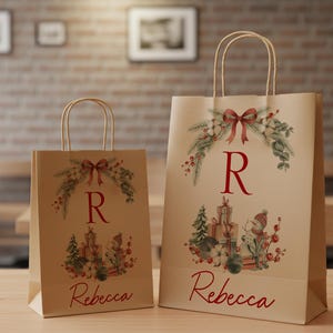 May include: Two kraft paper gift bags with festive watercolour designs. Each bag features a red bow, greenery, and the letter "R". The larger bag has the name "Rebecca" printed in red script. The bags are on a wooden table.