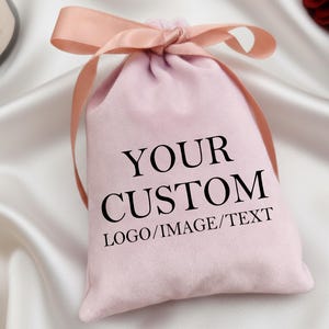 May include: A pale pink velvet drawstring bag with the text "YOUR CUSTOM LOGO/IMAGE/TEXT" in black. The bag is tied with a matching pink ribbon bow. The background is white satin.