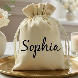 May include: A beige drawstring gift bag with the name "Sophia" in black script. The bag is on a gold-colored tray, with a lit candle and a vase of white roses in the background. The bag is made of a soft fabric.