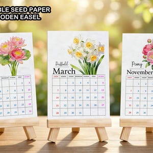 May include: Three plantable seed paper calendar cards displayed on small wooden easels. Each card features a floral illustration, the month name, and a grid. The text reads: PLANTABLE SEED PAPER FREE WOODEN EASEL, Water Lily July, Daffodil March, Peony November.