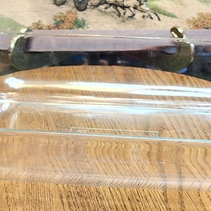 May include: Clear, oval-shaped glass baking dish with curved sides and handles. The dish is empty and sits on a wooden surface. The glass is transparent, allowing a view of the wooden surface below. The dish is designed for baking and serving.