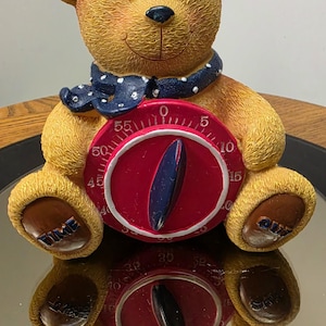 May include: A textured golden-brown bear figurine featuring a dark blue scarf with white polka dots. The bear holds a circular red kitchen timer with white numbers from 0 to 55 and a dark blue central dial. The word TIME is on the left foot and OUT is on the right foot.