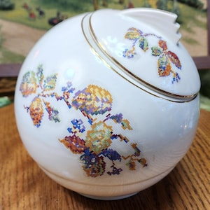 May include: A white ceramic jar with a lid, featuring a gold rim and colorful cross-stitch floral designs. The jar has a round shape with a small handle on the lid. The floral pattern includes shades of yellow, orange, blue, and green.