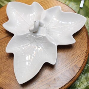 May include: A white, leaf-shaped serving dish with three sections. The dish has embossed leaf vein details and a small bird-like handle. It sits on a round, wooden serving board. The dish is likely used for serving snacks or appetizers.