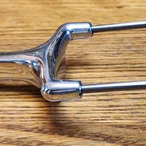 May include: A shiny, chrome-plated carving fork with two tines and a handle. The fork is designed for easy handling and is likely used for serving meat. The object is set against a wooden background.