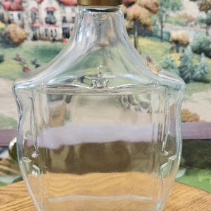 May include: A clear glass perfume bottle with a gold-colored cap. The bottle has a unique shape with a wide base and a narrower top. The background features a painting of a landscape with houses and trees.