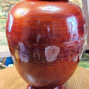 May include: A large, glossy, wooden vase with a lid. The vase is a deep reddish-brown color with visible wood grain. It has a rounded body, a pedestal base, and a small, pointed finial on the lid. The surface reflects light.