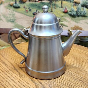 May include: A white ceramic mug with a black handle and the text "But First, Coffee" in a bold, black font. The mug is set against a blurred background, suggesting a cozy setting.