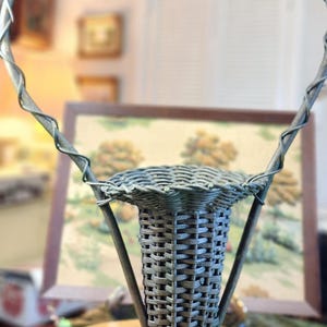 May include: A gray woven basket with a handle. The basket has a flared base and a cylindrical body. The handle is made of twisted metal. The basket is sitting on a wooden table. The background is blurred.