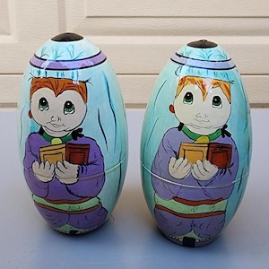 May include: Two hand-painted, egg-shaped nesting dolls. Each doll features a cartoonish figure with red hair, holding a book. The dolls are painted in shades of purple, blue, and yellow, with a black top.