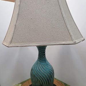 May include: A table lamp featuring a textured, teal-colored ceramic base with a swirling, wave-like pattern and a tapered, light beige fabric lampshade. The base sits on a clear, circular glass stand, and the lamp has a simple, functional design.