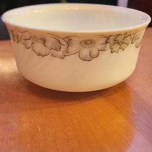 May include: Off-white ceramic bowl with a floral design in brown. The bowl is set on a wooden surface. The floral pattern is repeated around the rim of the bowl. The bowl is a kitchenware item.