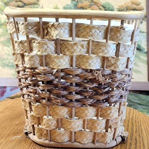 May include: A woven basket with a rounded top and a rectangular base. The basket is made of natural-colored woven materials, with alternating bands of light and dark brown. The basket is empty and appears to be handmade.