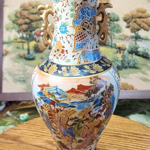 May include: A decorative ceramic vase with a flared rim and two gold-colored handles. The vase features detailed hand-painted scenes of figures and landscapes in shades of blue, red, and gold against a white background. The base and rim have floral patterns.