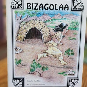 May include: A book titled "BIZAGOLAA" with the subtitle "Historical Apache Dolls to Color and Cut." The cover art depicts a running figure in traditional clothing near a dwelling. The book's border has decorative tribal patterns. The author is Jan Mike, and the artist is Cathie Lowmiller.