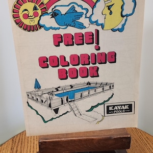 May include: A rectangular paper sign featuring a colorful illustration of a sun, a rainbow, a blue bird, and a crescent moon above the text FREE! COLORING BOOK. Below, a drawing of a swimming pool with a deck and the Kayak Pools logo.