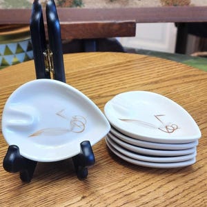 May include: A set of six white ceramic ashtrays with a leaf-like shape and a gold and brown design. The ashtrays are stacked on a wooden surface, with one displayed on a black stand. The ashtrays are a vintage style.
