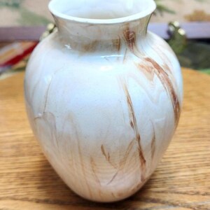 May include: A small, off-white vase with brown marbling. The vase has a rounded body and a slightly flared rim. The decorative vase is likely made of glass or ceramic and would be a nice addition to any home decor.
