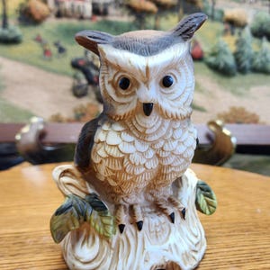 May include: A ceramic owl figurine with detailed feather textures, perched on a tree stump with green leaves. The owl has large, dark eyes and a brown and cream color scheme. The figurine is a decorative item.