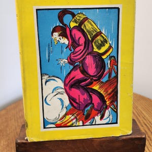 May include: A vintage book titled The Campfire Girls as Detectives with a bright yellow cover. The central illustration depicts a figure in a pink jumpsuit and yellow backpack leaping from a red structure amidst blue clouds and rain.